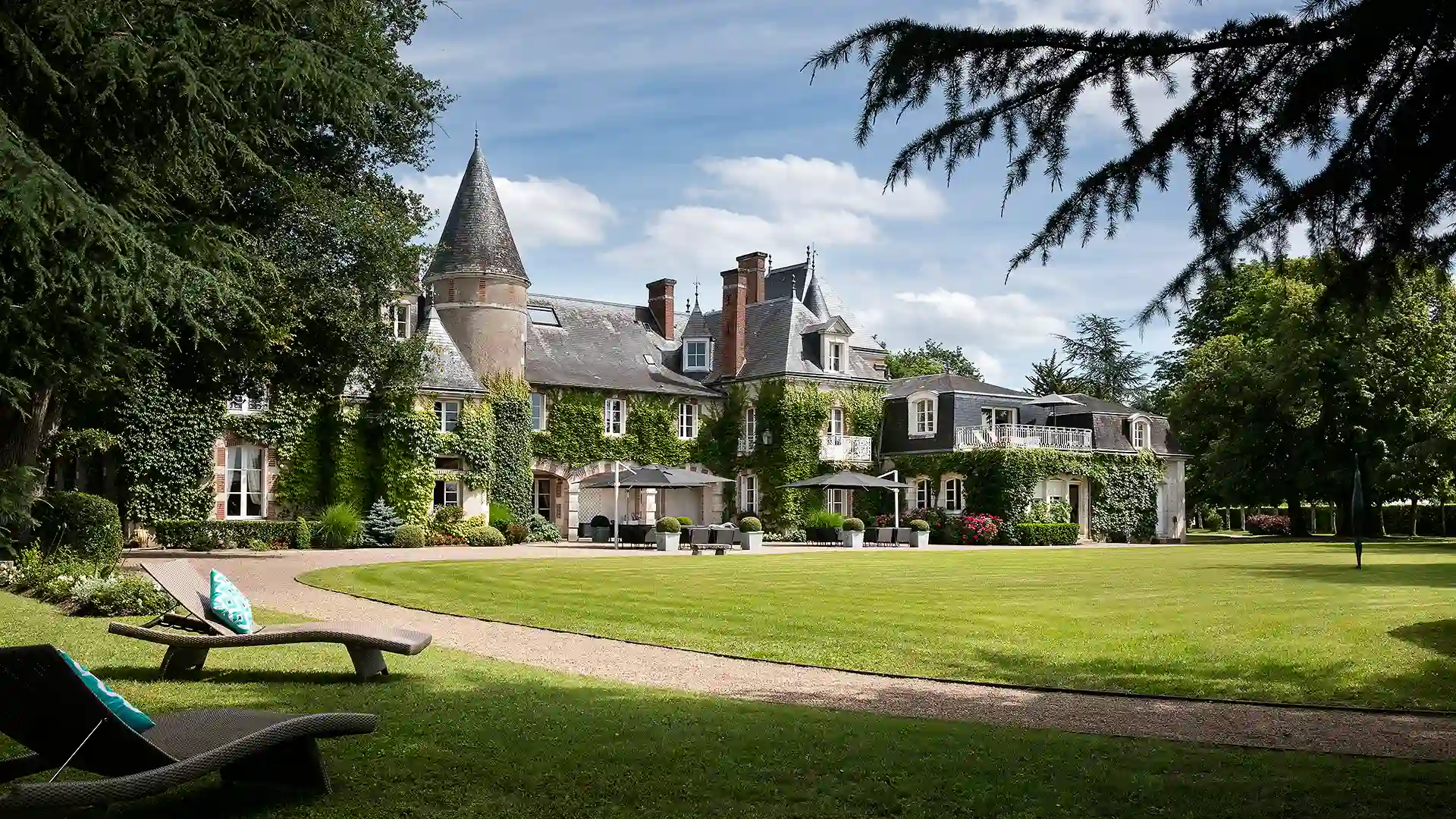 Totale des Hauptgebäudes des Schlosses unter sonnigem Himmel, mit Grünflächen und Liegestühlen. Hier befindet sich das Hotel Les Hauts de Loire – ein 5-Sterne-Hotel mit Restaurant und Spa.