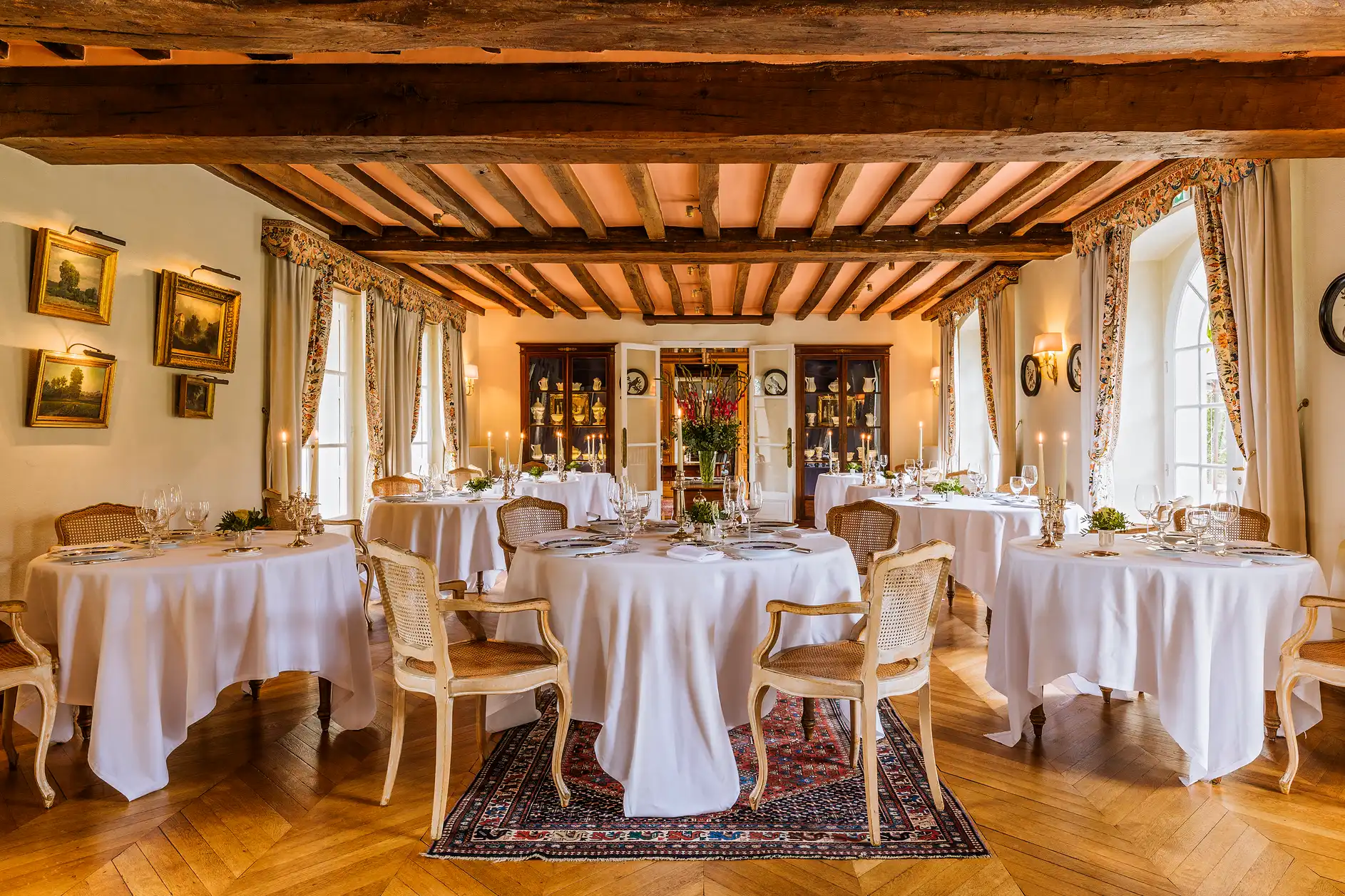 Salle de restaurant élégante avec tables rondes nappées, parquet et charpente en bois au plafond.