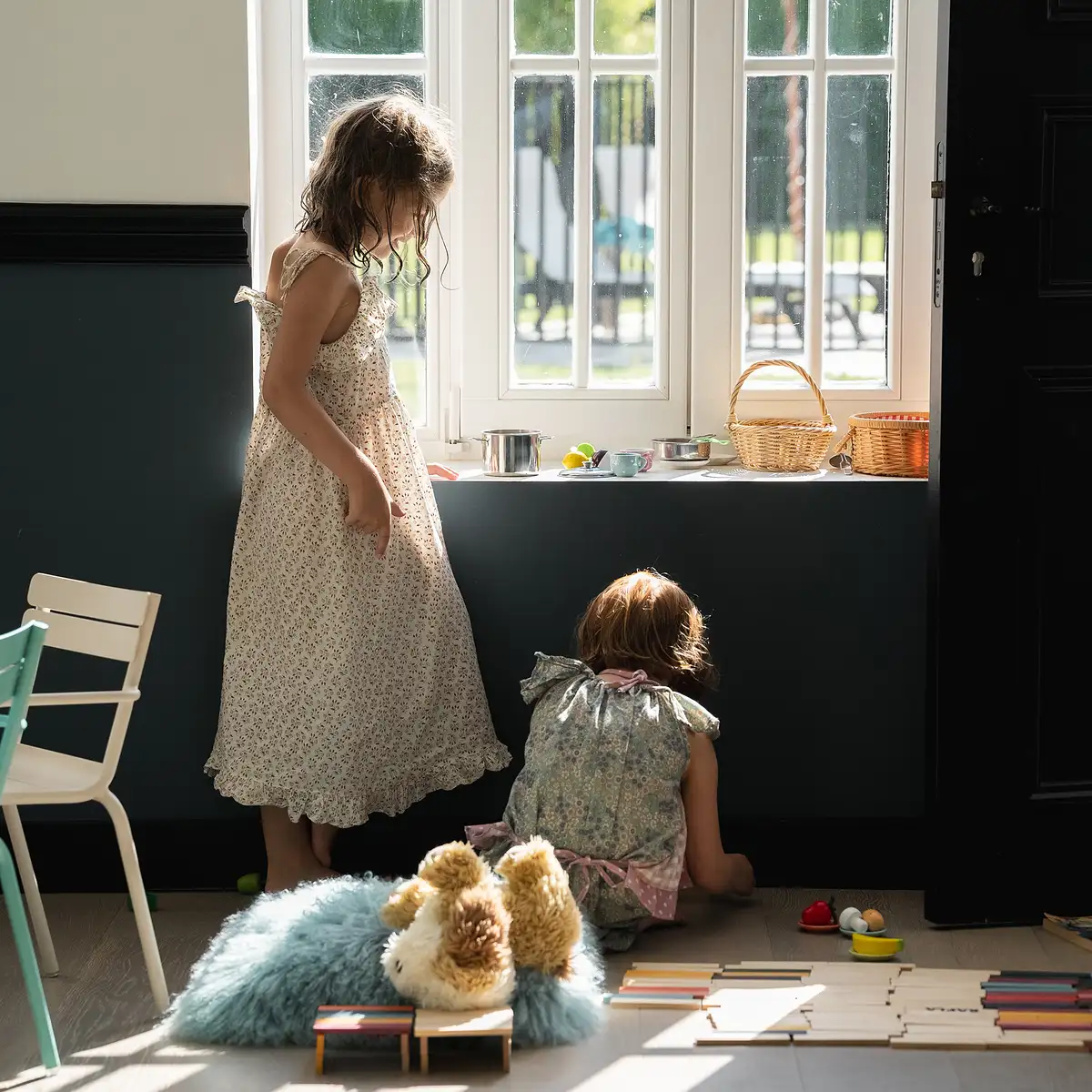 Zwei Kinder schauen aus einem großen Fenster in einem Spielzimmer mit grauen und weißen Wänden.