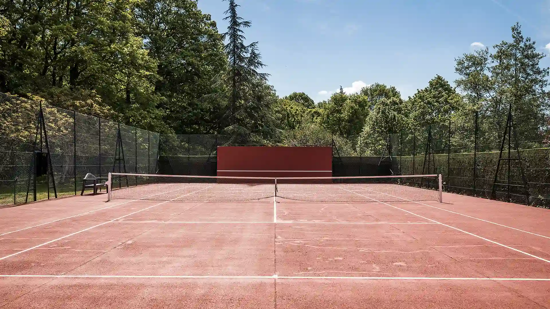 Perspective latérale du court de tennis en terre battue, montrant la clôture grillagée et la rangée d'arbres en bordure. Les Hauts de Loire – Hôtel, Restaurant & Spa – 5 étoiles.