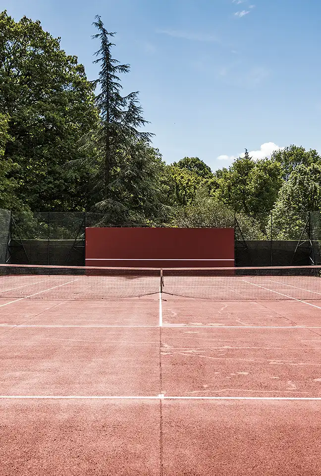 Medium shot of the clay tennis court, highlighting the net and the red wall in the distance. Les Hauts de Loire – Hotel, Restaurant & Spa – 5-star.