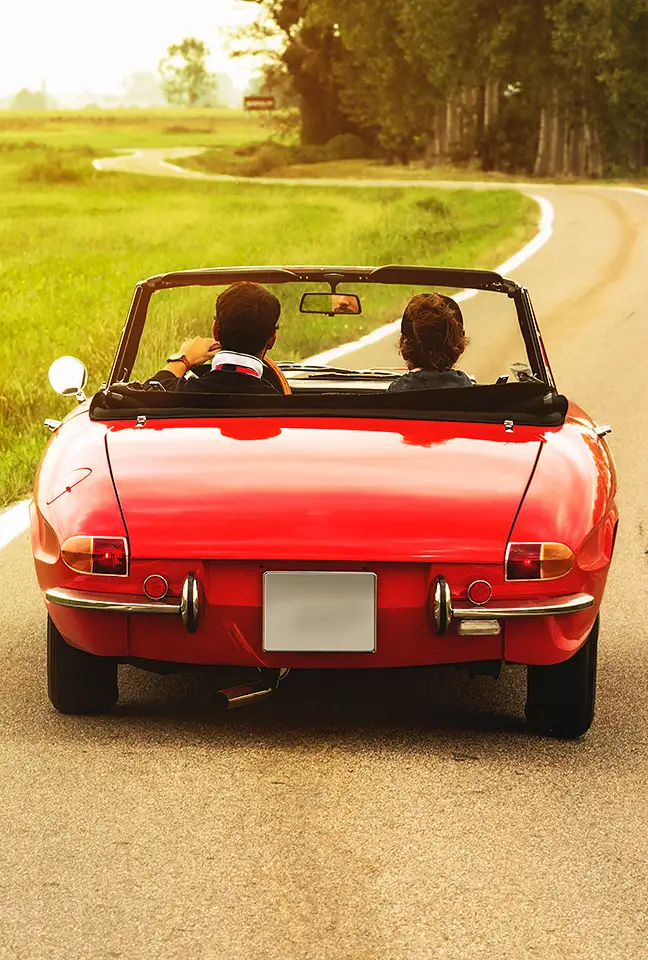 Una pareja conduce un descapotable rojo por una carretera pequeña y pintoresca en una tarde soleada.