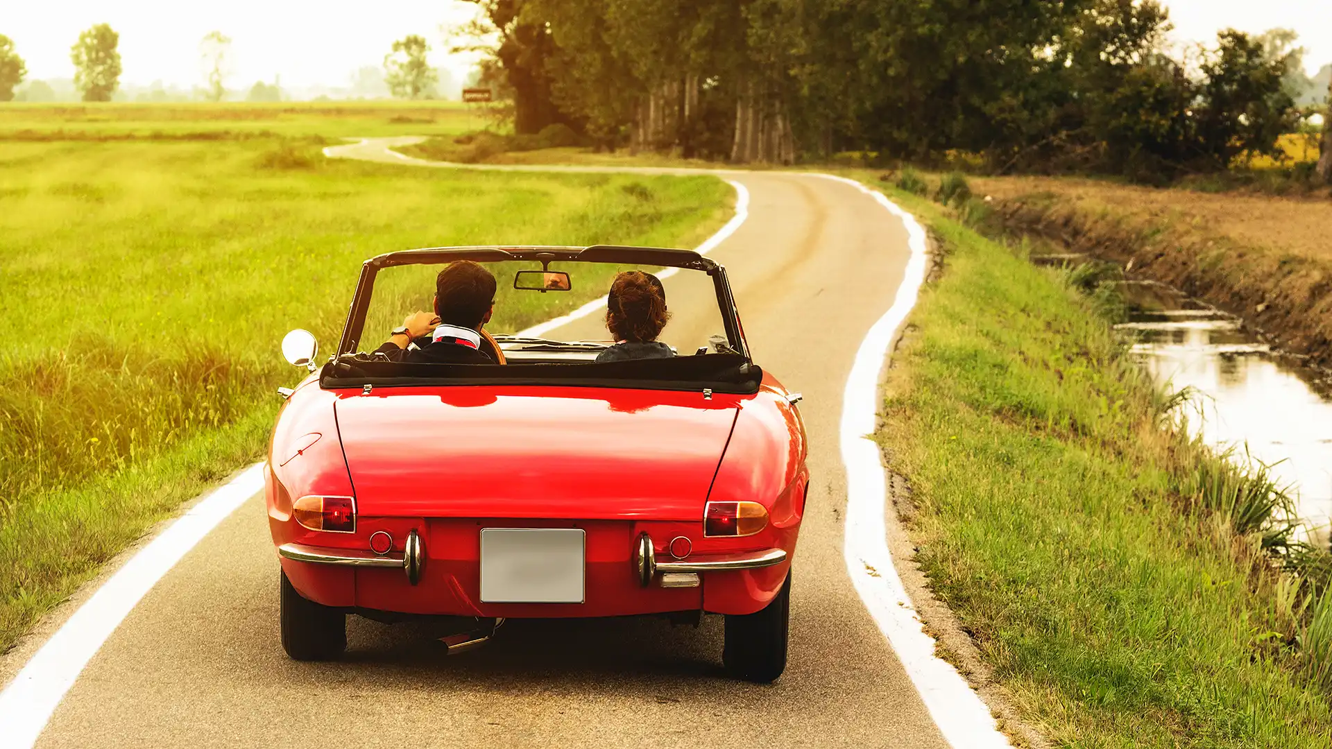 Red vintage car, viewed from behind, with its two occupants out for a drive on a road at sunset.