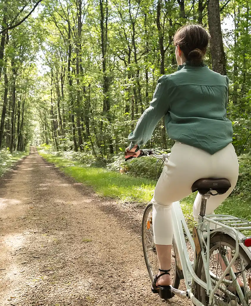 Vue de dos d'une femme en tenue claire roulant à vélo sur un chemin forestier ensoleillé. Une activité proposée par Les Hauts de Loire – Hôtel, Restaurant & Spa – 5 étoiles.