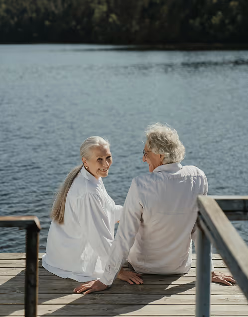 Senior couple sitting on a wooden dock by the water, holding hands and smiling at each other.