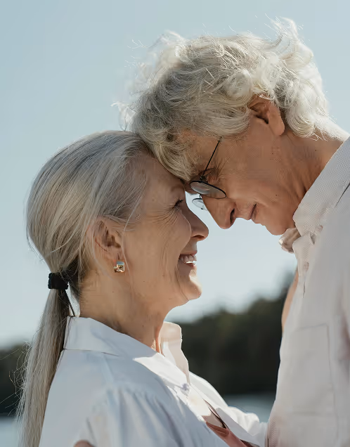 Elderly couple with foreheads touching, smiling warmly at each other outdoors.