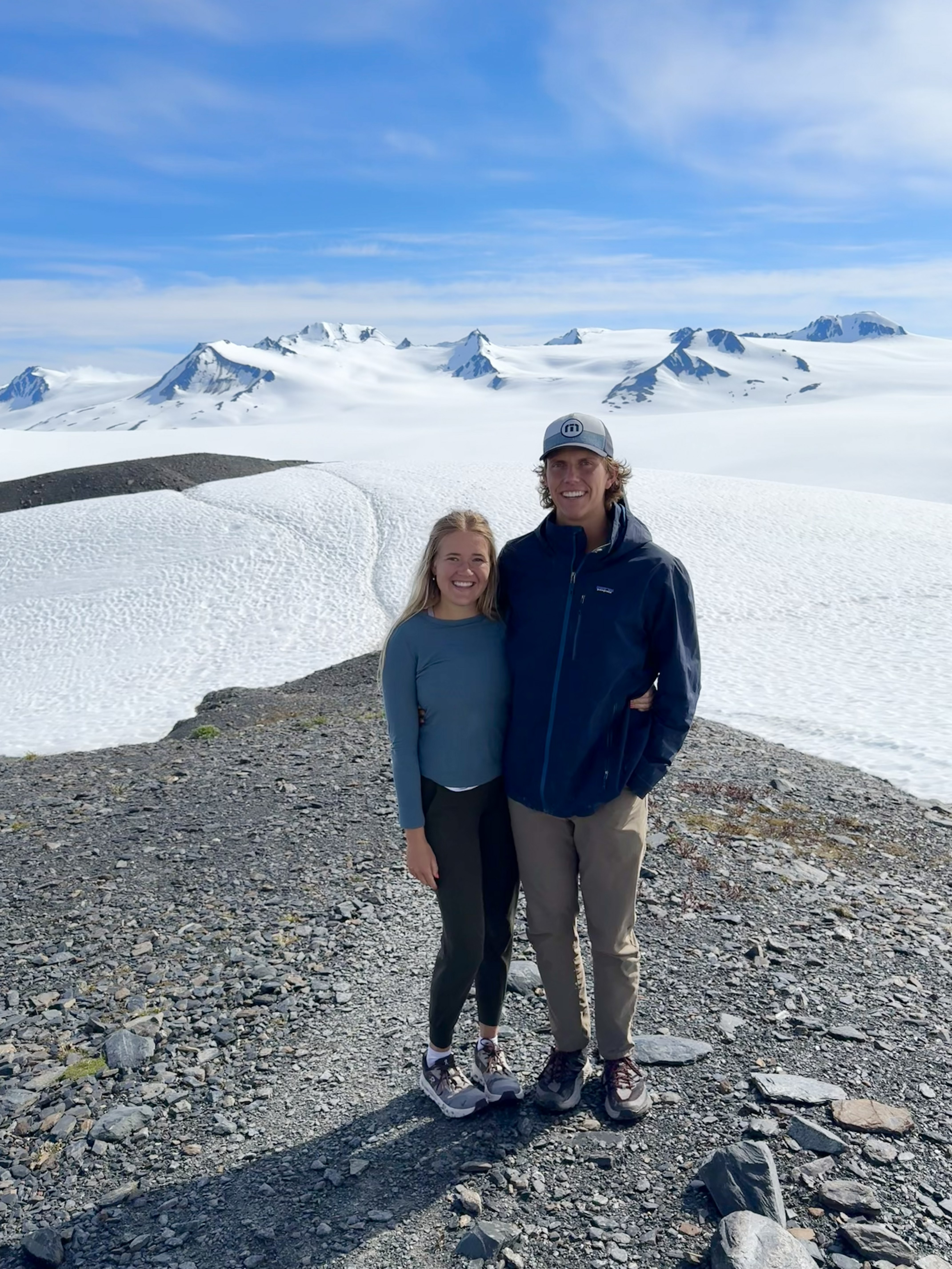 Smiling couple in outdoor clothes standing on rocky ground with snowy mountains in the background.