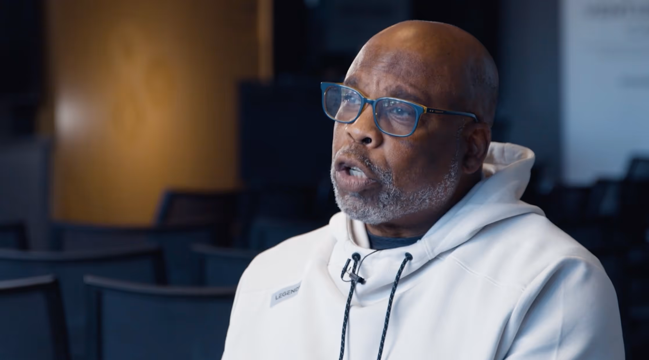 Man with glasses and gray beard speaking while wearing a white hoodie in a dimly lit room.