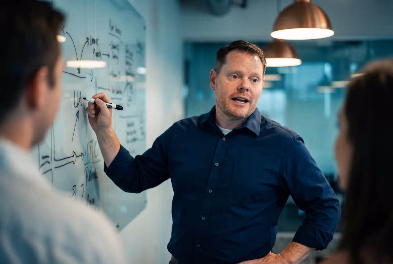 Man explaining ideas on a glass board while two colleagues listen attentively in an office.