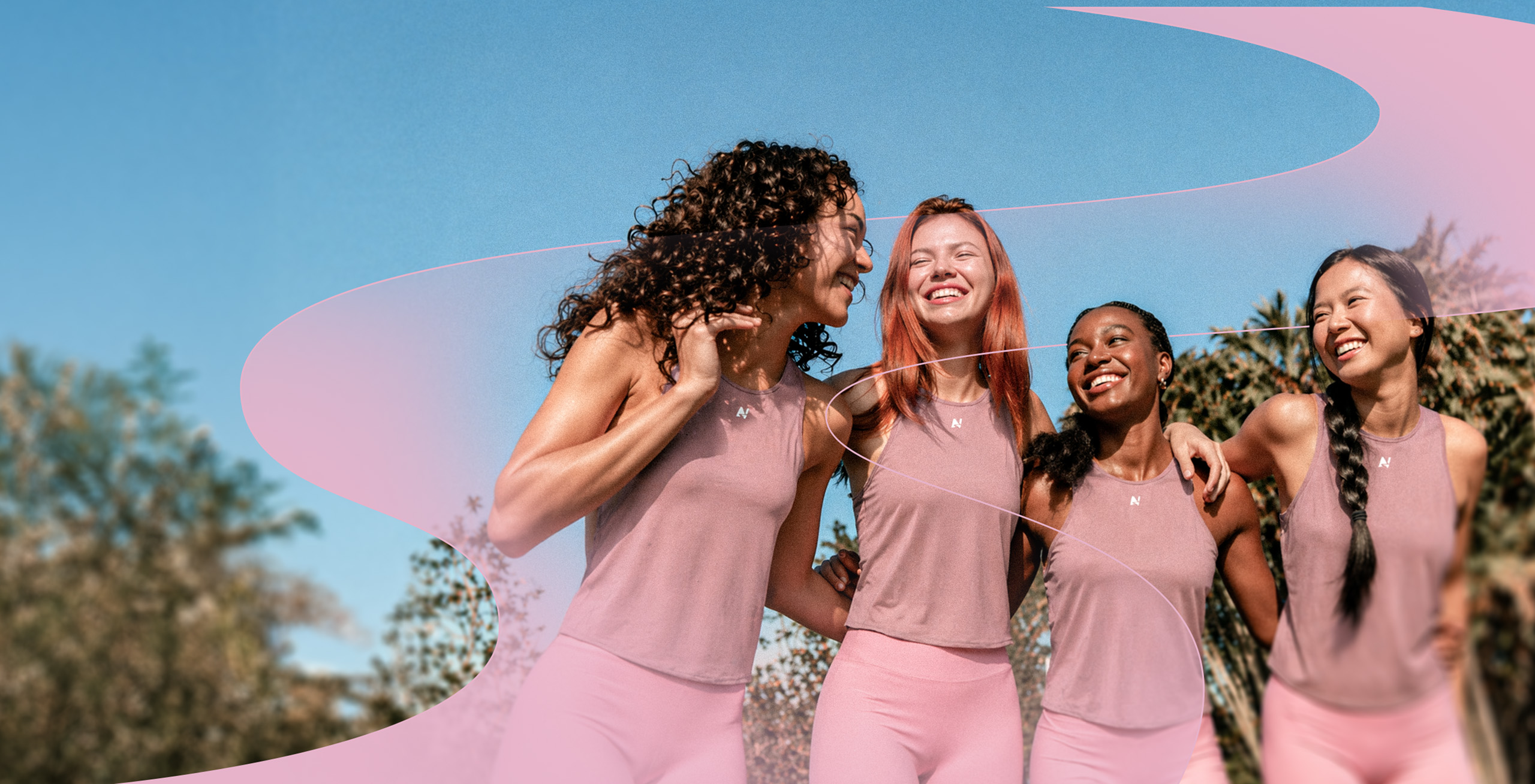 Four smiling diverse women in pink workout clothes outdoors with blue sky and trees behind them.