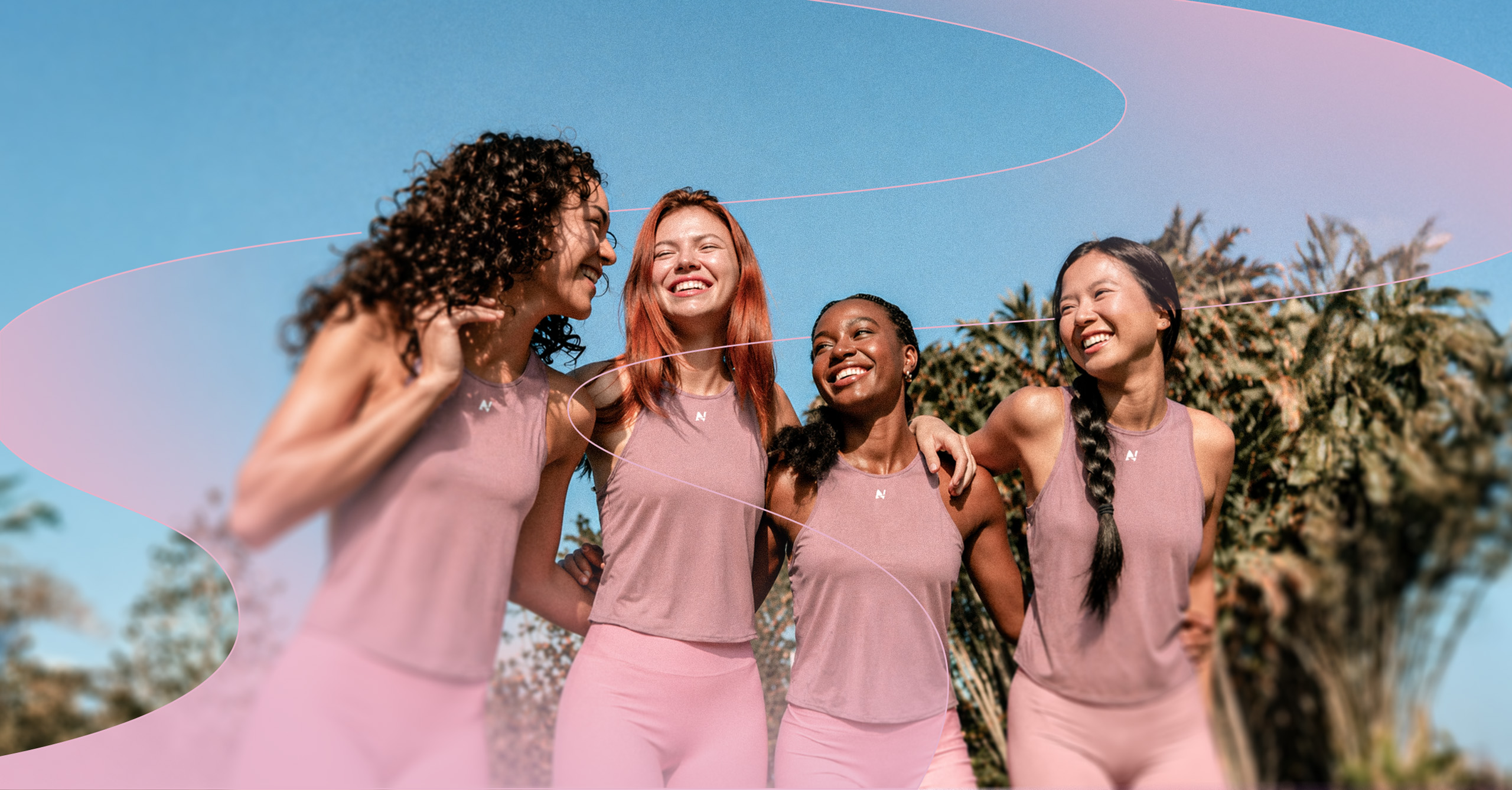 Four smiling women in pink activewear with arms around each other outdoors under blue sky.