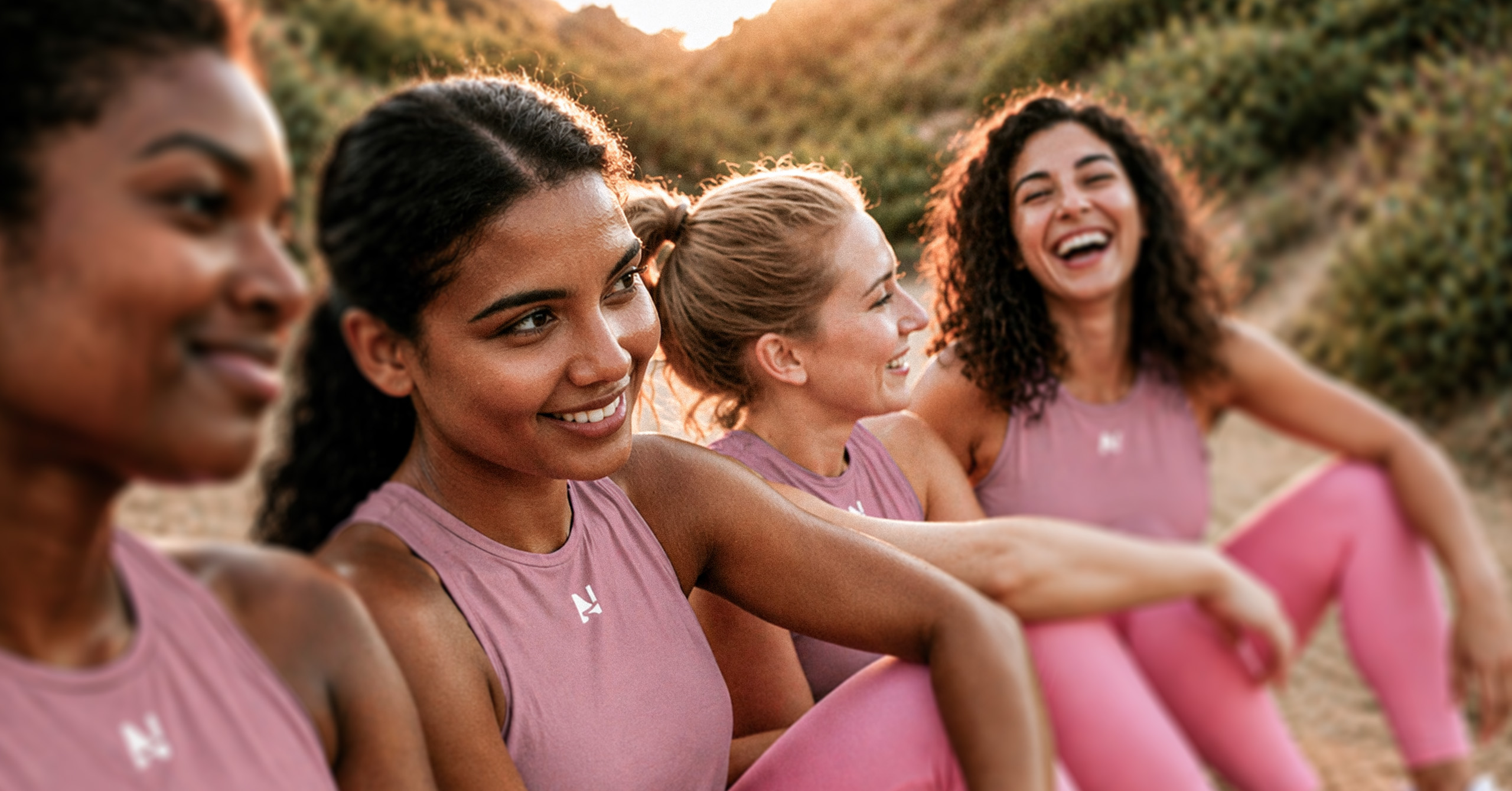 Four diverse women in lavender workout tops and pink leggings sitting outdoors, smiling and laughing.
