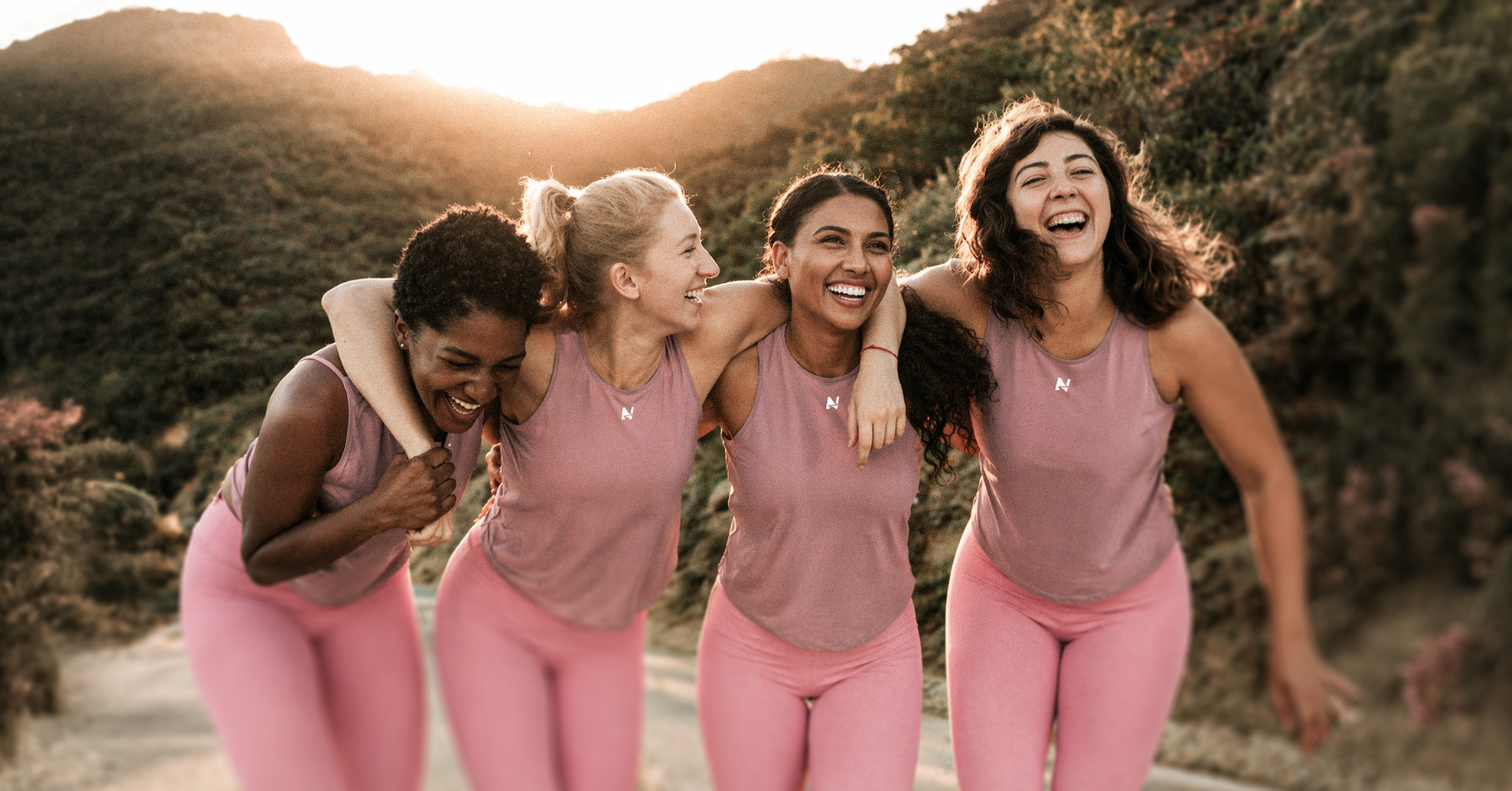 Four women in pink outfits laughing and embracing outdoors on a sunny day near hills.