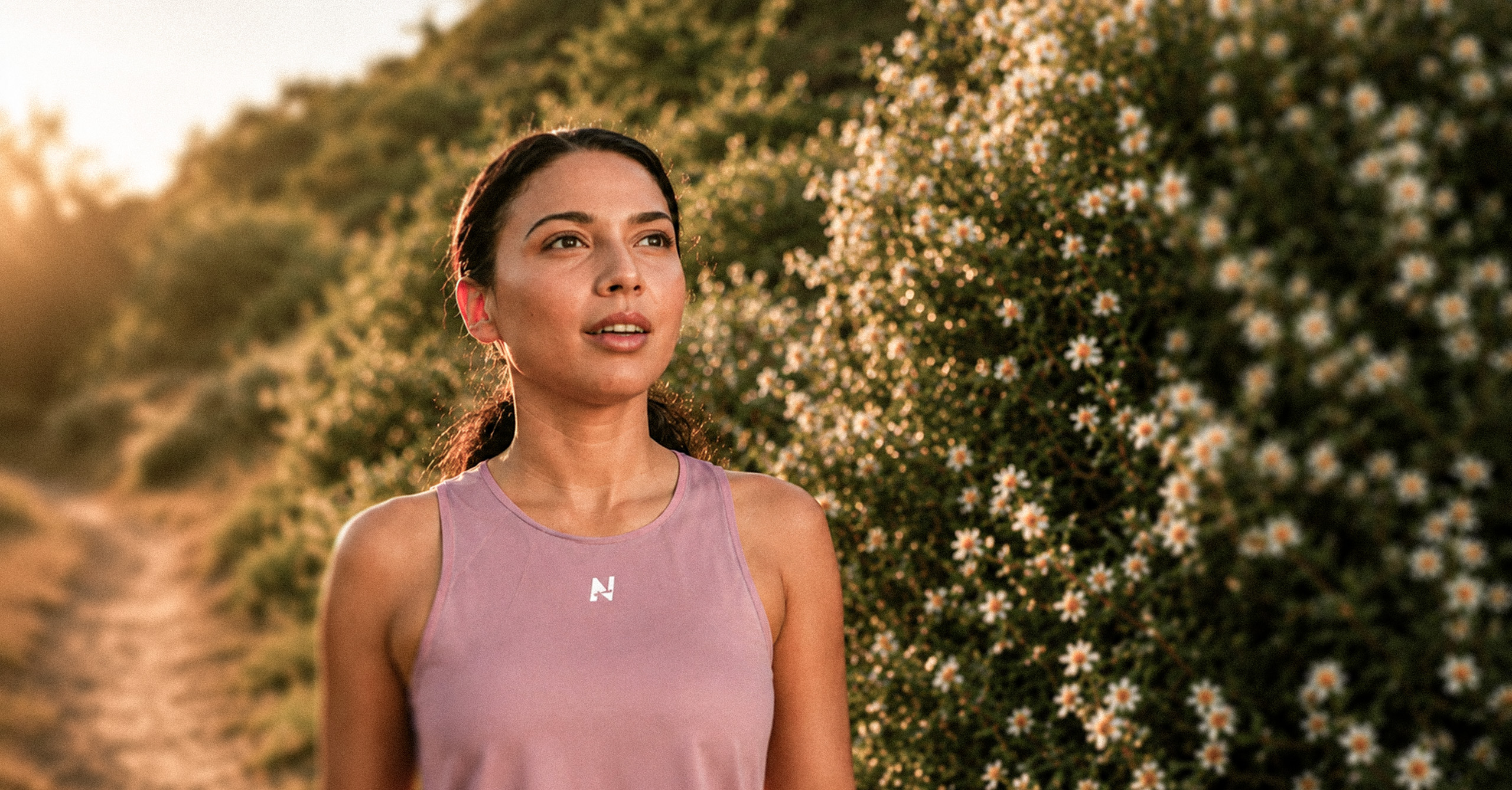 Young woman in pink tank top standing near a flowering bush on a sunny outdoor path.