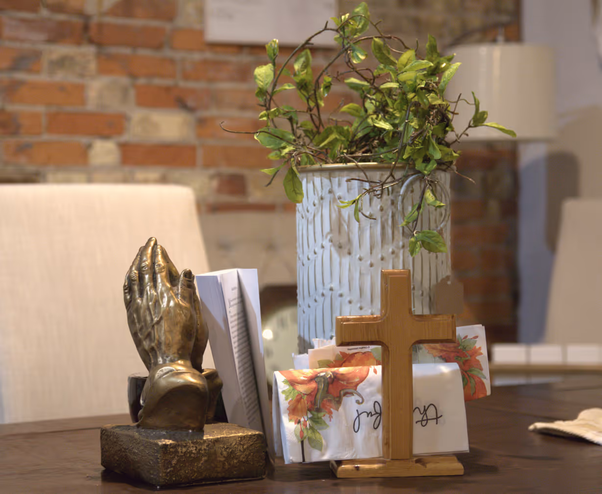 Table display with praying hands statue, wooden cross, napkins, and potted green plant against brick wall.
