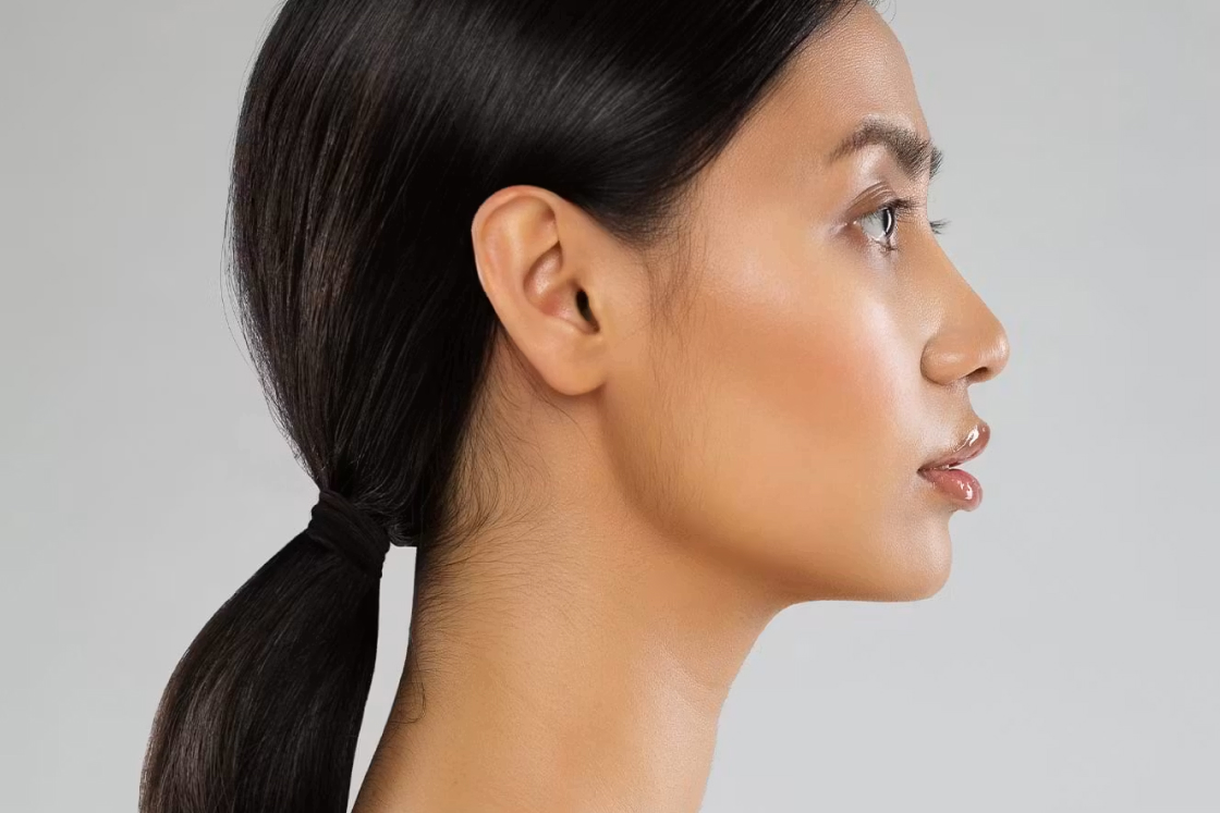Profile of a woman with smooth skin and dark hair tied back in a ponytail against a gray background.