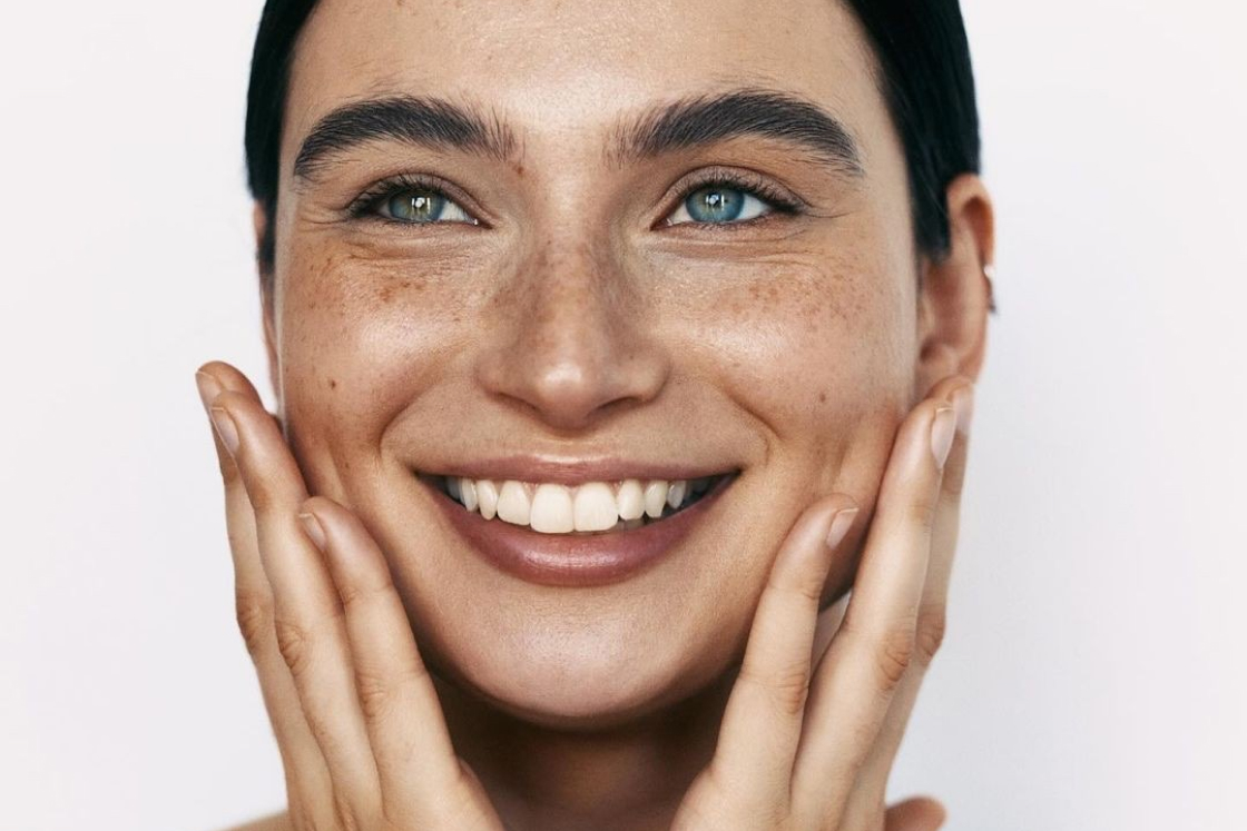 Close-up of a smiling woman with freckles touching her cheeks and looking to the side.