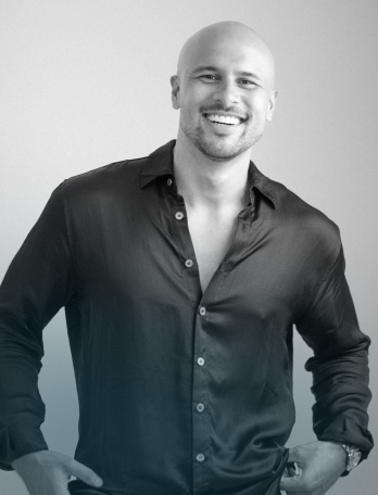 Smiling bald man in a dark buttoned shirt with hands on hips against light background.