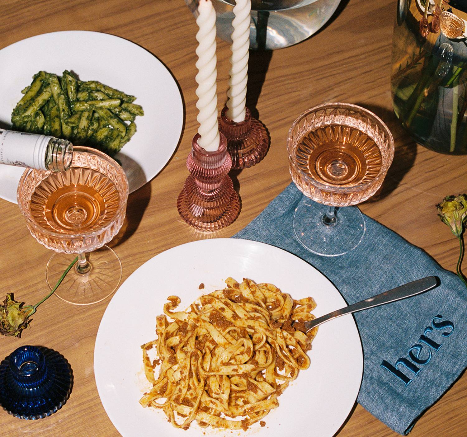 A dinner setting with pasta, pink glassware, candles, and blue linen napkin