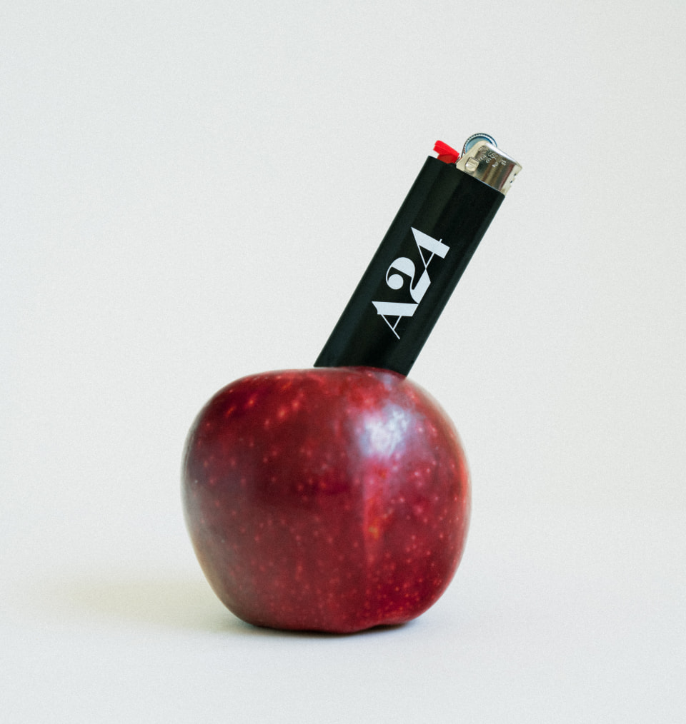 Black A24 Films lighter coming out of a rerd apple next to branded black tote bag with double handles, next to close up shot of Netflix black tumbler with red logo