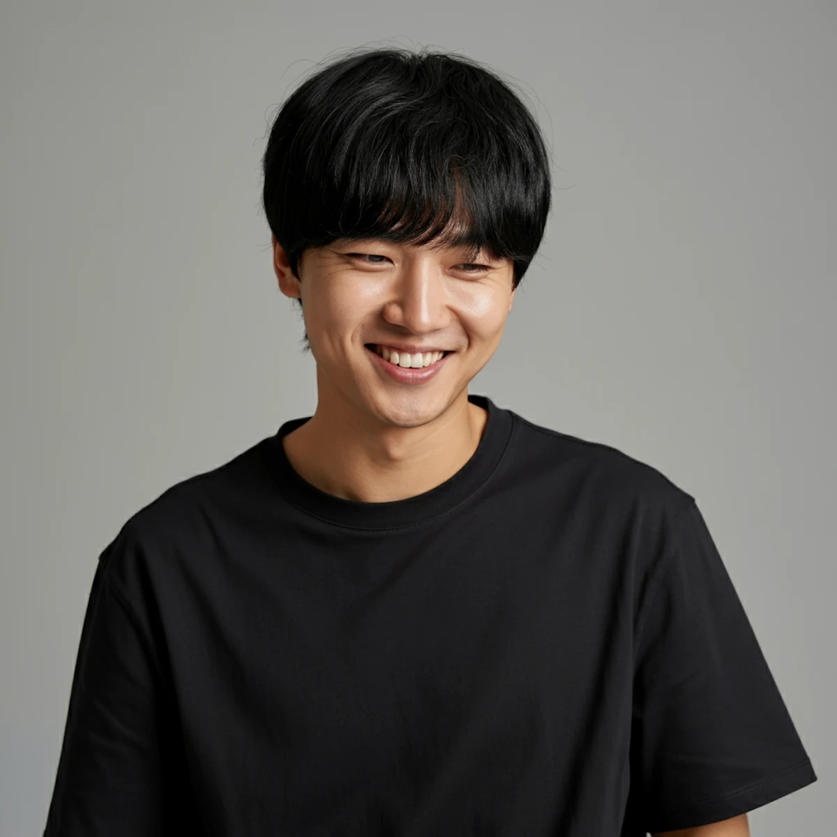 Young man with black hair smiling and looking down, wearing a plain black t-shirt against a gray background.