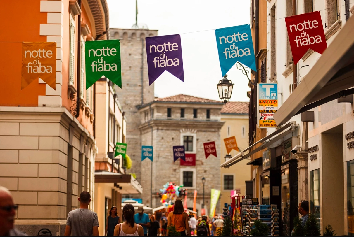 Bandiere colorate con scritto 'notte di fiaba' lungo una strada a Riva del Garda, con la Rocca sullo sfondo