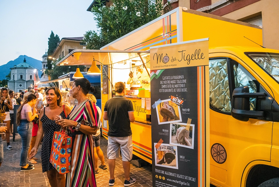 Persone in fila davanti a un food truck giallo che serve tigelle in una serata all'aperto.