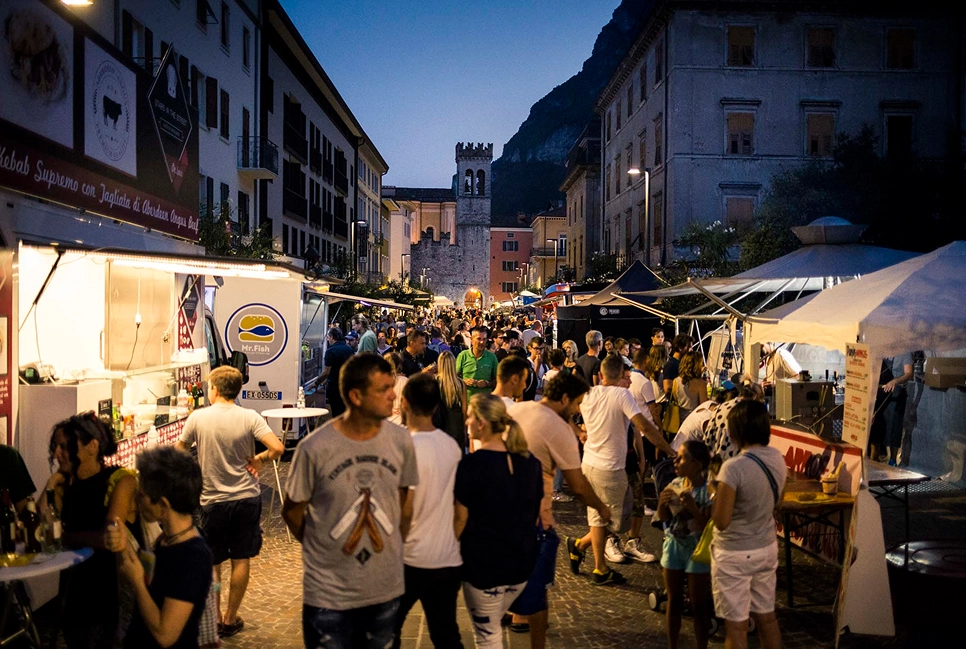 Folla in una via affollata di un mercato serale con bancarelle e edifici storici sullo sfondo.