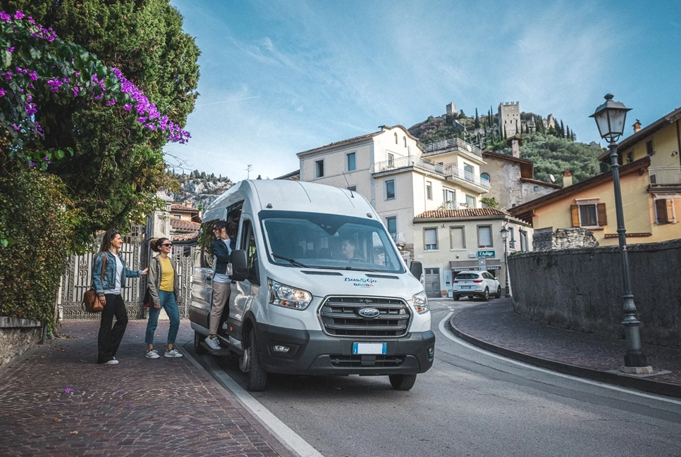 Persone che salgono su un van bianco parcheggiato in una strada di paese con colline e castello sullo sfondo.