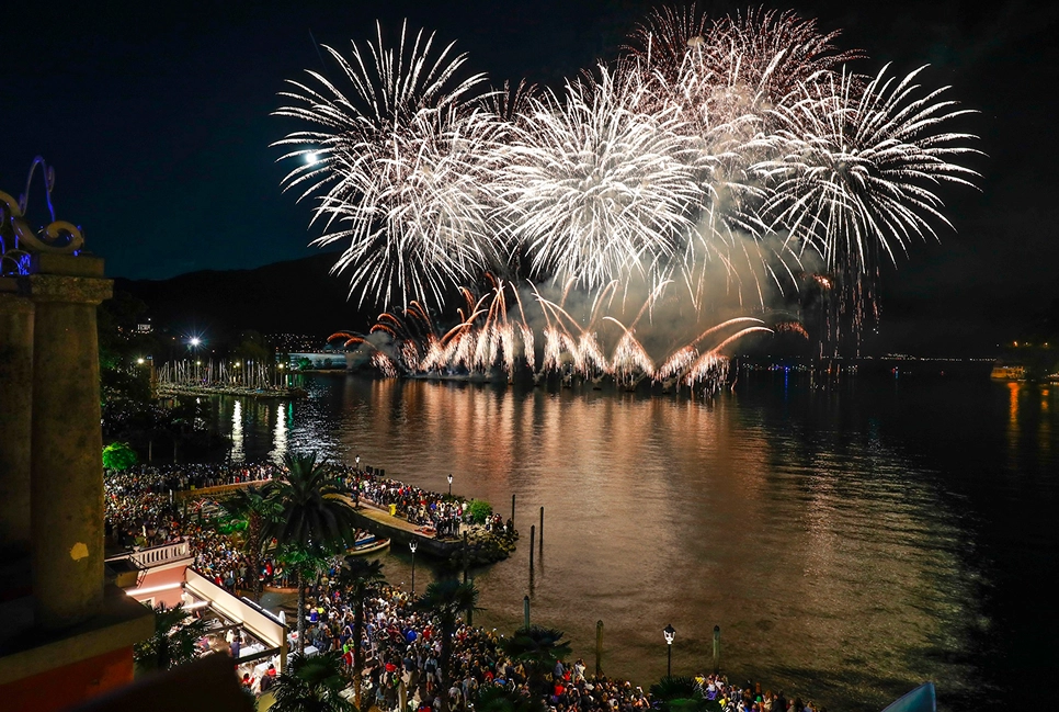 Fuochi d'artificio bianchi sul lago con una folla radunata lungo la riva di notte.