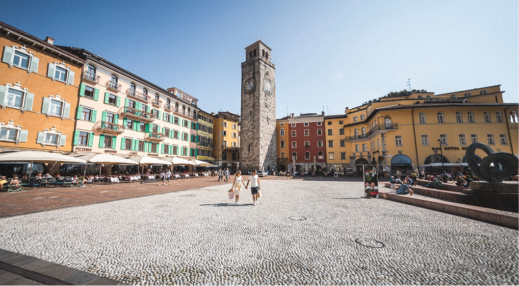 Piazza con Torre Apponale e edifici colorati con caffè all'aperto, persone camminano e siedono.