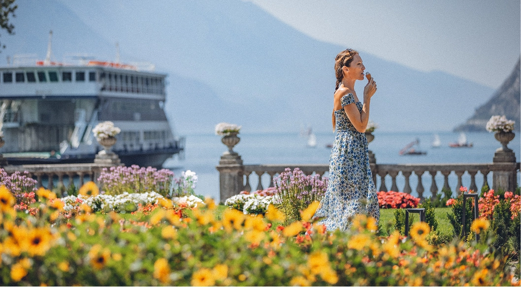 Donna con abito floreale che mangia un gelato in un giardino fiorito con vista lago e barca.