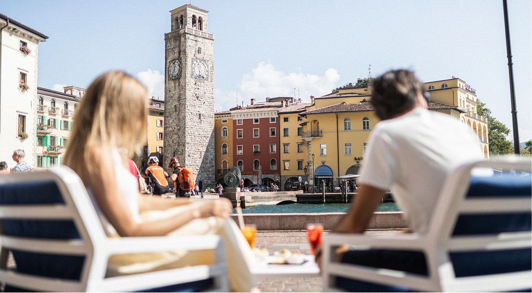 Una coppia seduta su sedie all'aperto con vista su torre Apponale e edifici colorati sul lungolago.