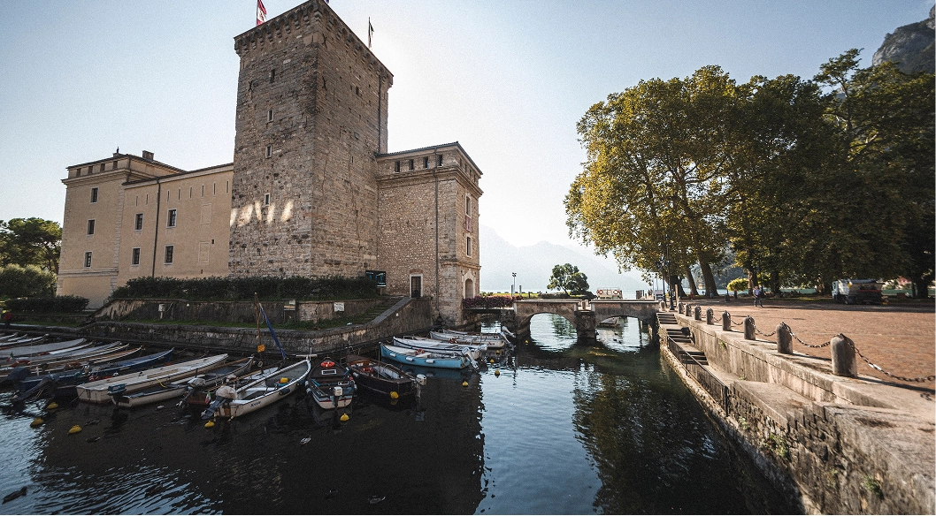 Rocca di Riva del Garda con barche attraccate sul lago e alberi lungo il camminamento al sole.