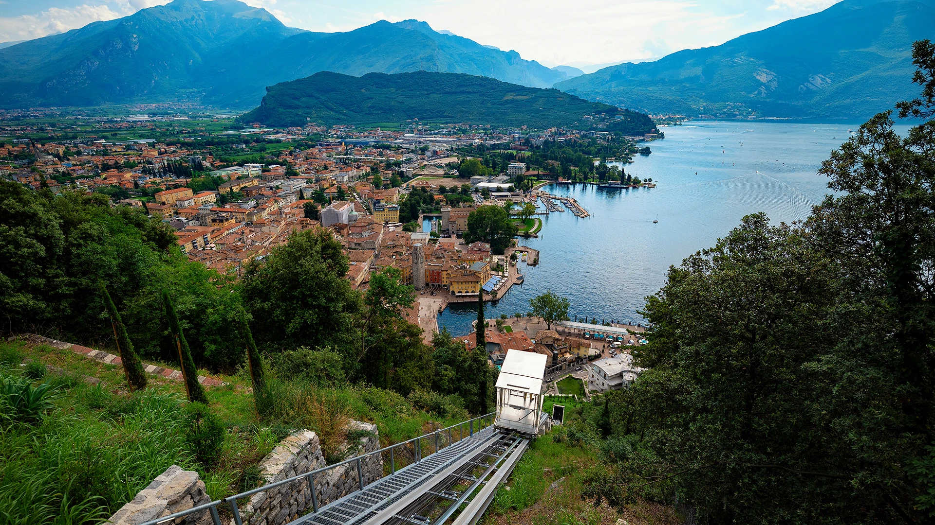 Vista panoramica di un lago con città, montagne e una funicolare in discesa nella natura verde.