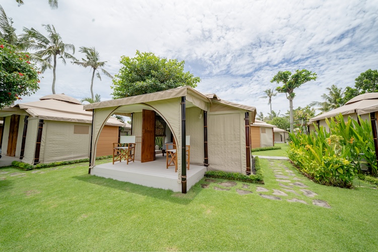 Outdoor glamping tents with wooden furniture on a wooden deck surrounded by lawn and tropical plants under a cloudy sky.