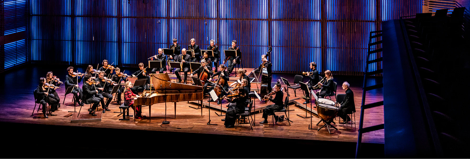 Orchestra performing on stage with musicians playing strings, piano, brass, and percussion instruments.