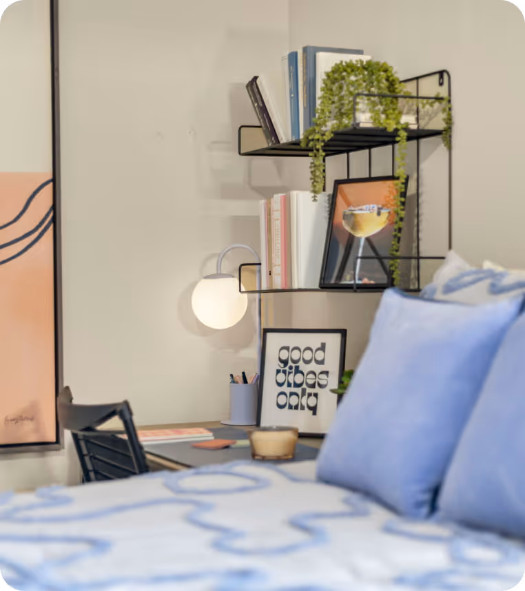 Cozy bedroom corner with a blue bedspread, framed 'good vibes only' sign, small desk with lamp, and wall shelves holding books and a plant.