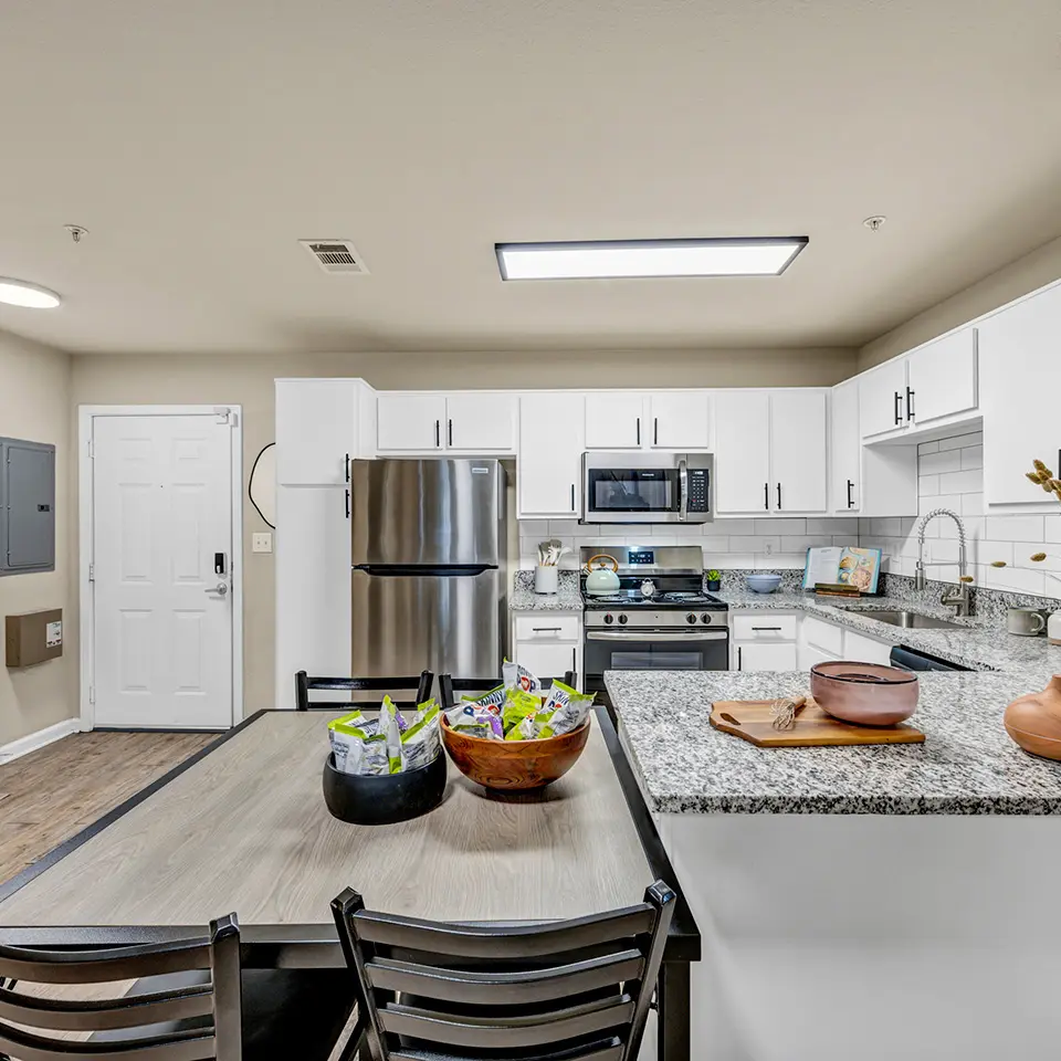 Modern kitchen with stainless steel refrigerator, stove, microwave, white cabinets, granite countertops, and a wooden dining table with bowls of snacks.