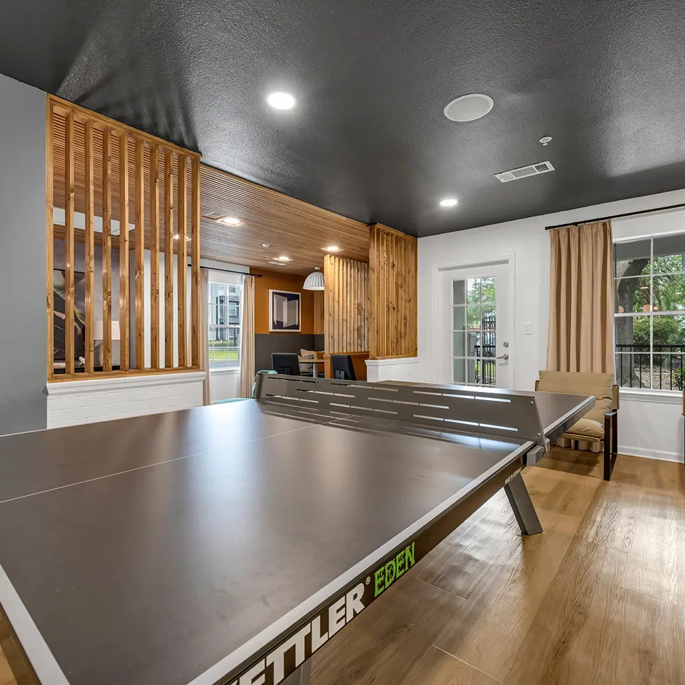 Indoor recreation room with a close-up view of a gray Kettler Eden ping pong table, wooden floor, and seating area with beige curtains and modern decor.