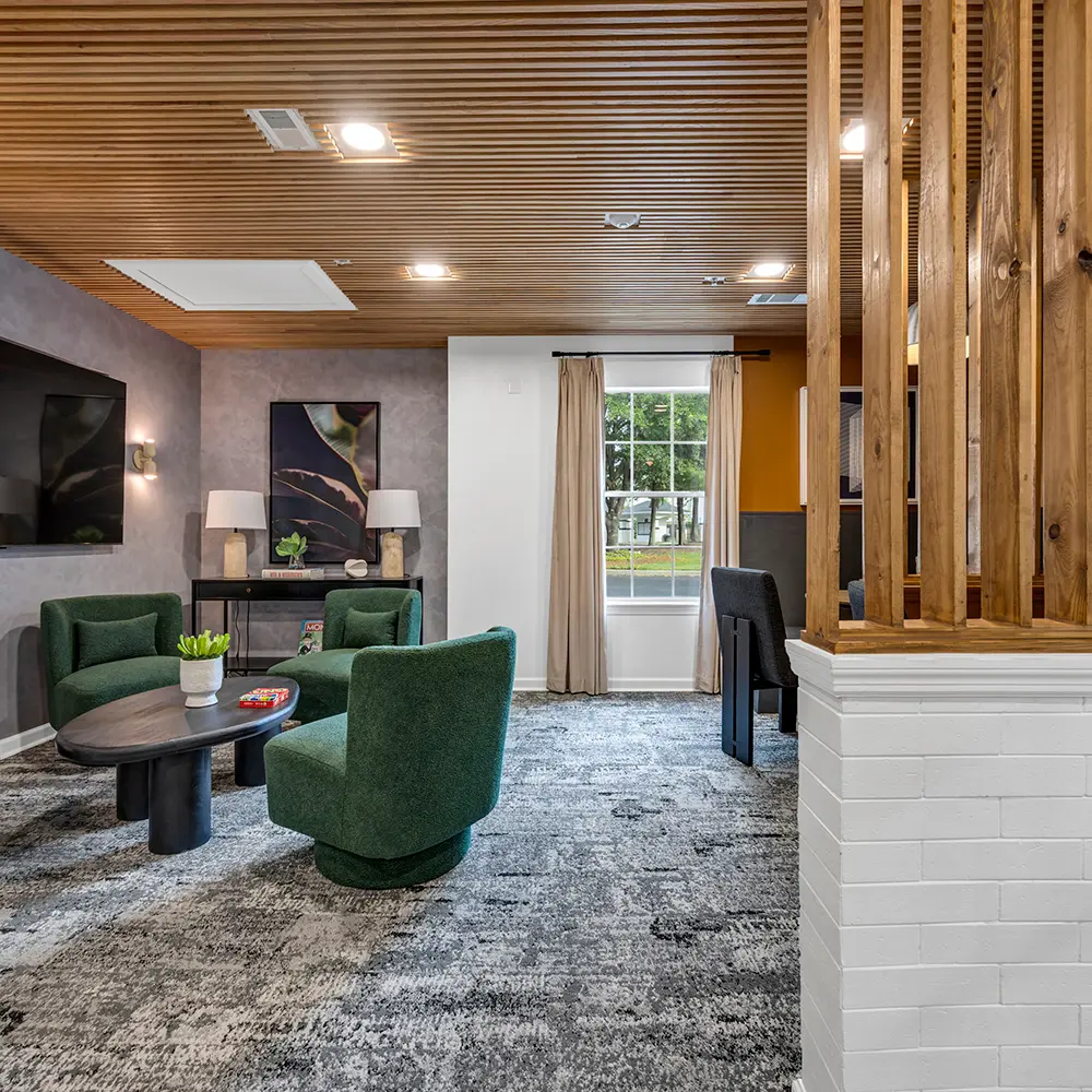 Modern lounge area with three green upholstered chairs around a black oval coffee table, large TV on wall, and a window with beige curtains.