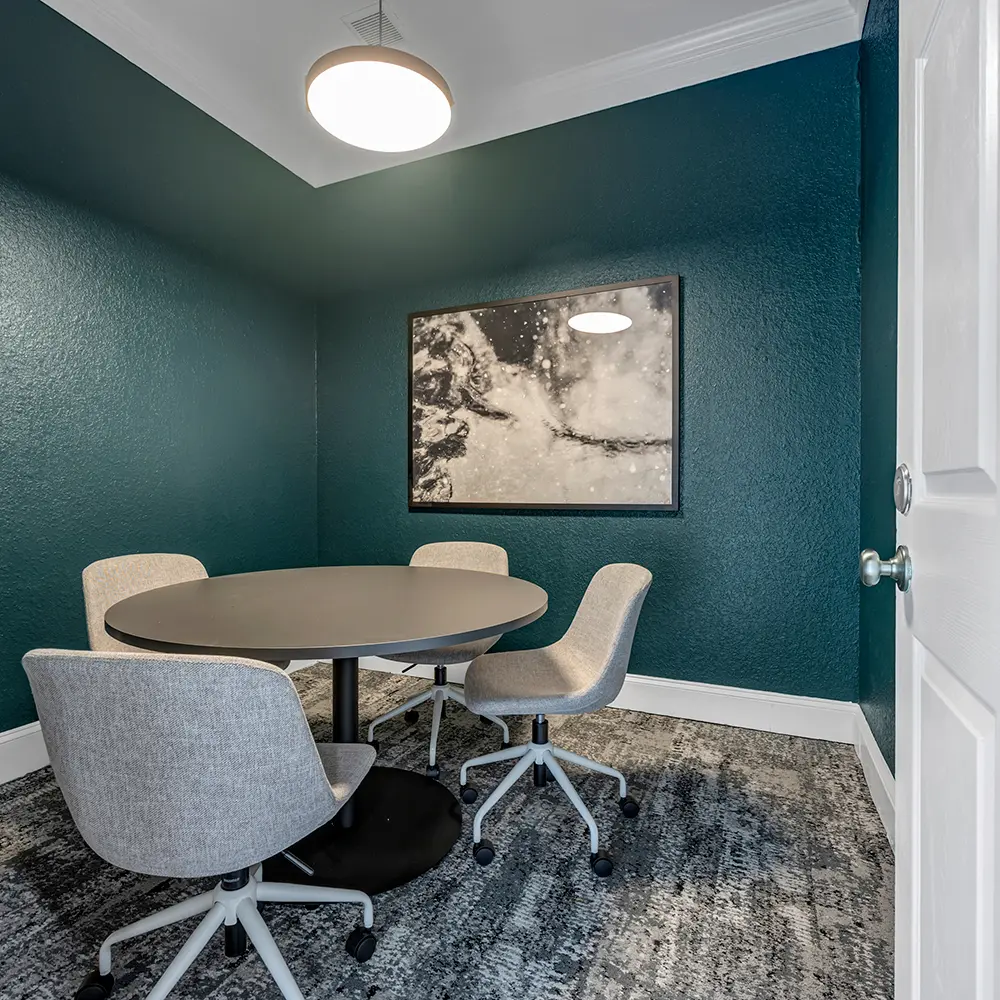 Modern study room with teal walls, round grey table, four grey swivel chairs, abstract wall art, and overhead circular light.