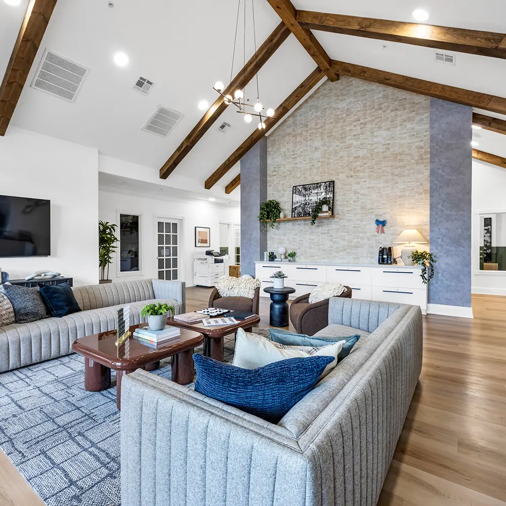 Modern living room with vaulted ceiling and wooden beams, gray sectional sofa, two brown armchairs, and coffee tables on a patterned rug.
