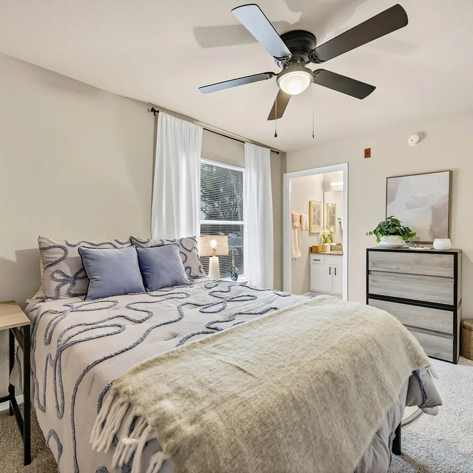 Neatly made bed with patterned comforter and two blue pillows in a bright bedroom with ceiling fan.
