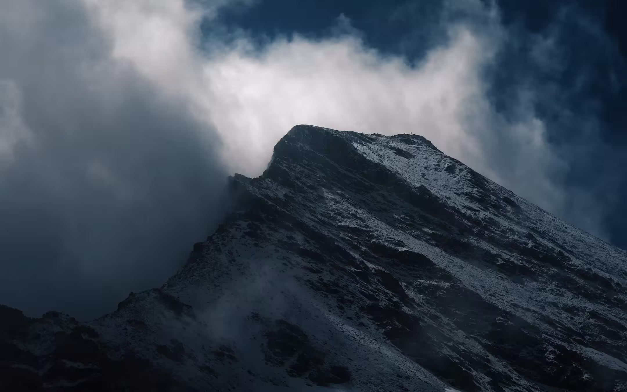Snow-covered mountain peak with dark rocky slopes under moody cloudy sky.