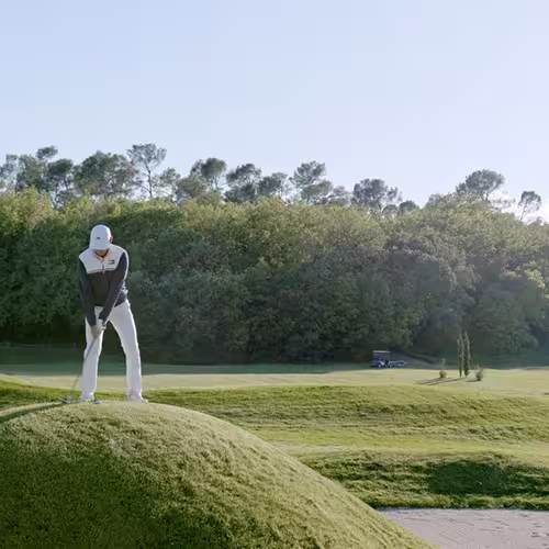 Golfer preparing to swing on grassy mound with trees and clear sky in the background.