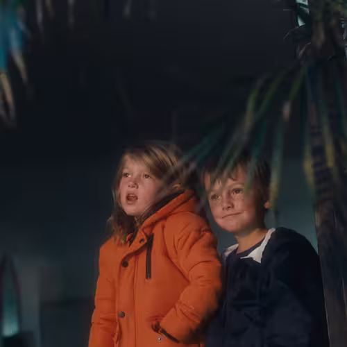 Two children, one in an orange jacket, looking up with interest in a dark setting.