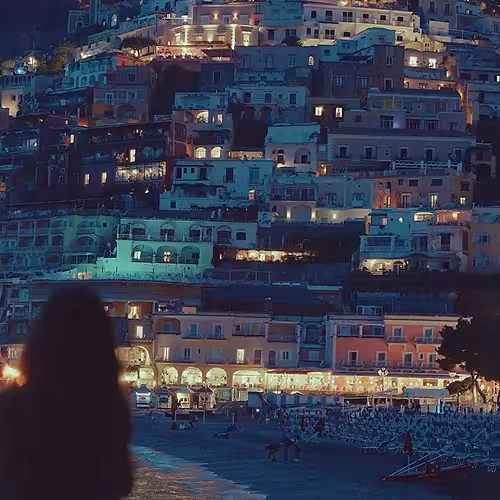 Silhouette of person looking at a lit hillside town with buildings cascading down to a beach at night.