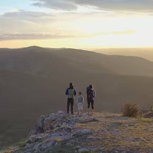 Three hikers stand on a rocky cliff overlooking a hazy mountain landscape at sunset.