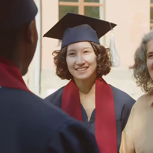 Smiling female graduate in cap and gown with red stole at outdoor ceremony with others.